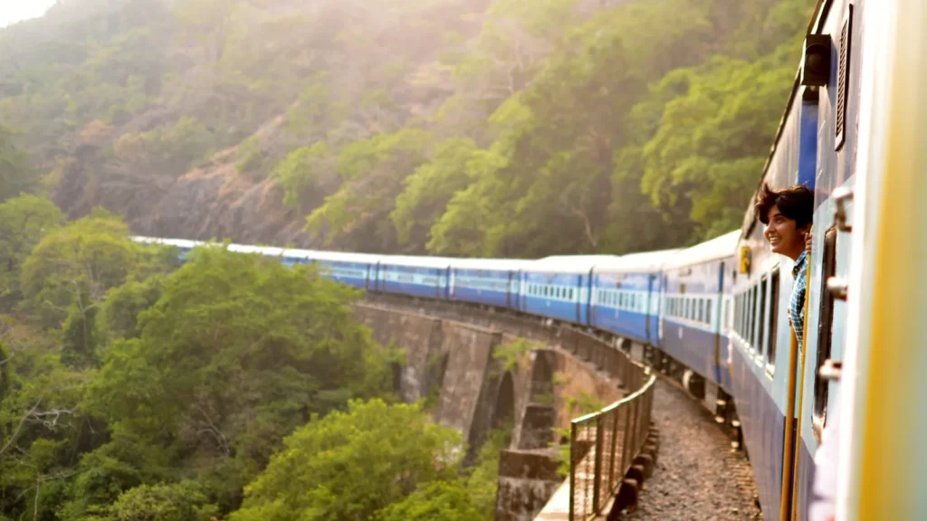 A long blue passenger train curving along a hillside track through green forest in soft daylight