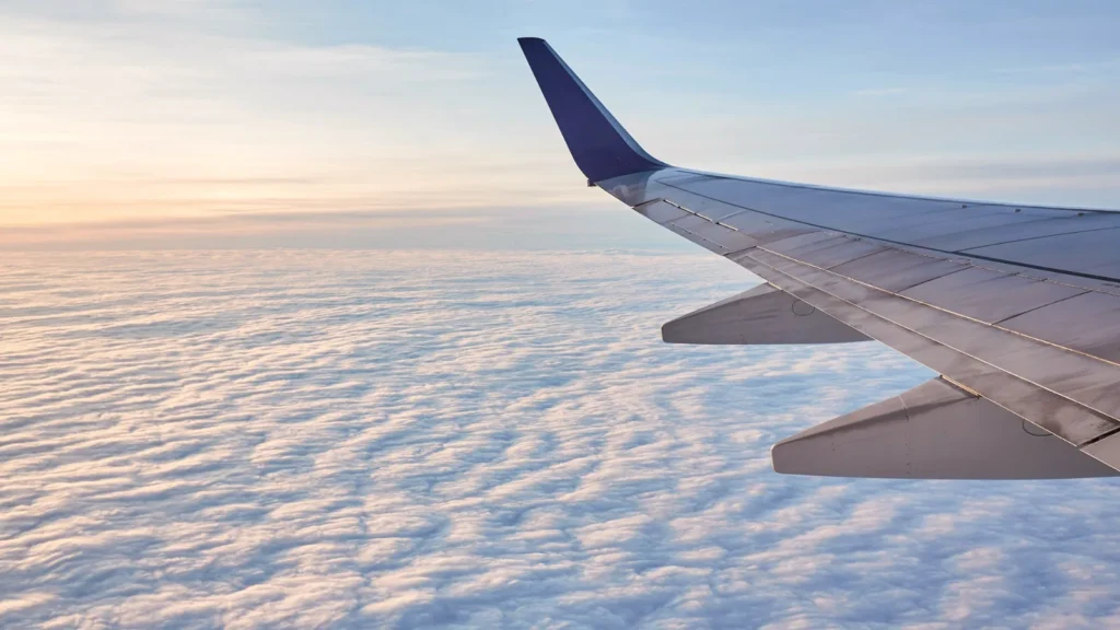 An aircraft wing seen from a window seat above a blanket of clouds in soft daylight